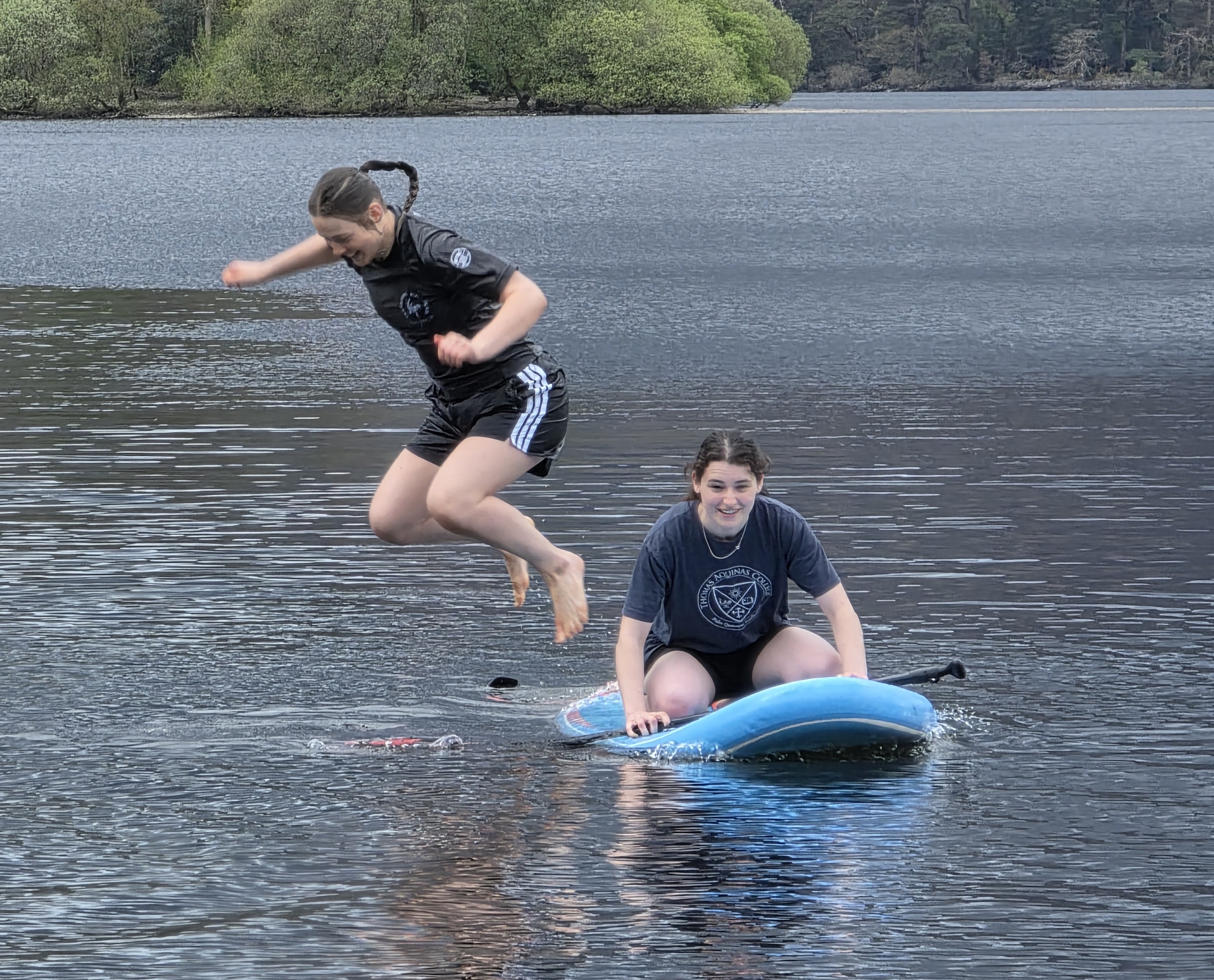 Two team members larking about on a paddleboard on the lake, one leaping into the water